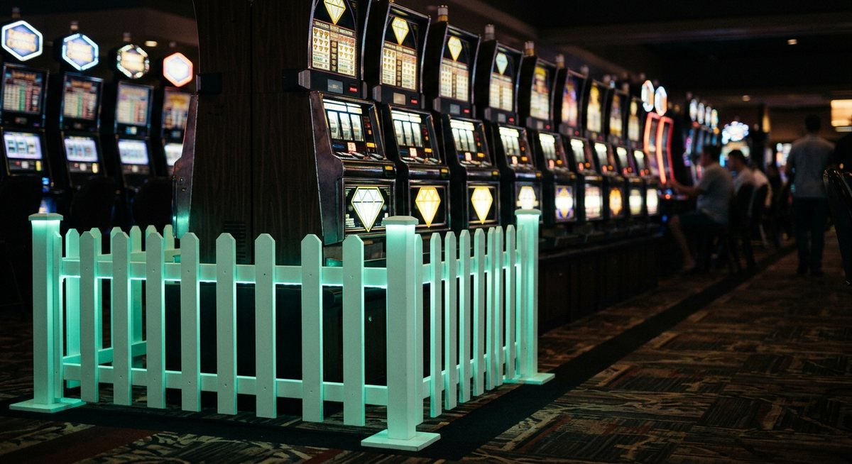 Pagar kecil bergaya neon mint berdiri di depan deretan kabinet slot gelap dengan simbol diamond bercahaya di kejauhan